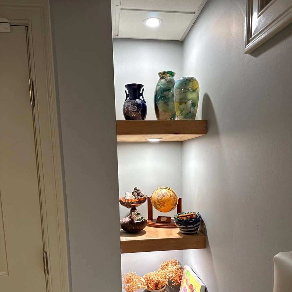Wood floating shelves in a narrow alcove with individual lights illuminating pottery and decorative objects